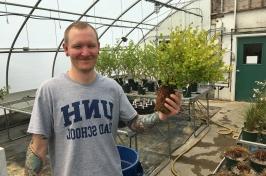 Male student in greenhouse holds a plant. His t-shirt says 主要研究 研究生院.